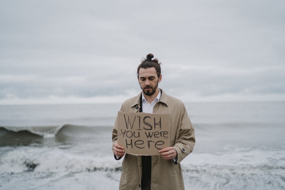 A man in a trench coat stands by the sea holding a 'Wish you were here' sign, expressing longing.